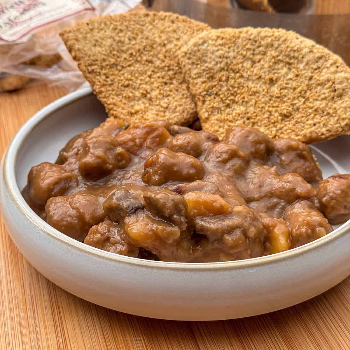 A bowl of thick Scottish stovies with visible chunks of meat and vegetables, served with two large, crispy wholegrain crackers on a wooden surface.