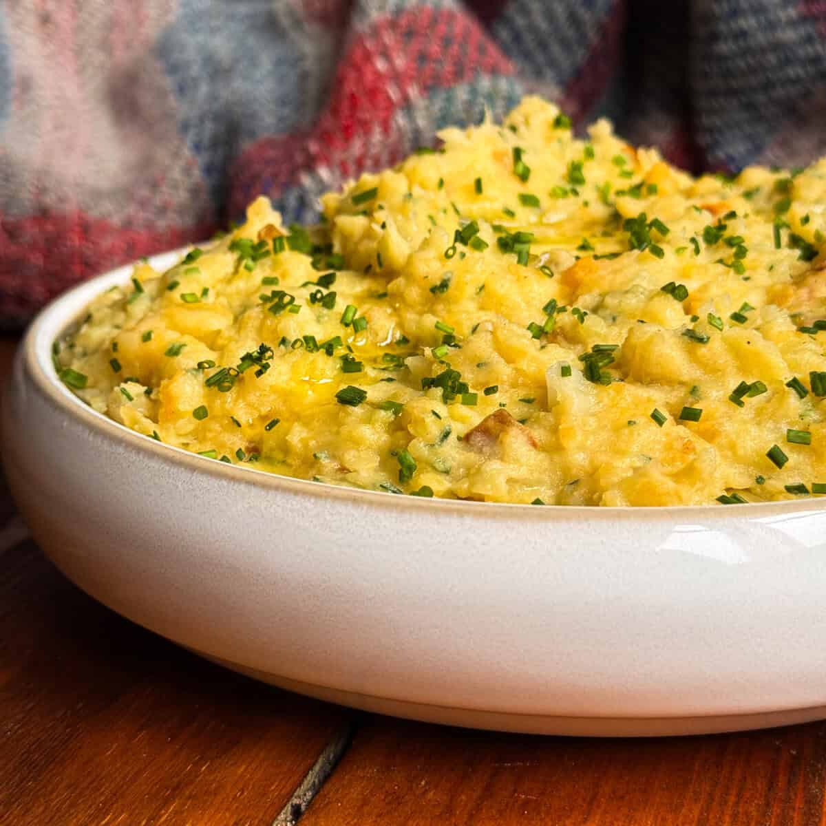 A close-up of a white dish filled with scottish clapshot topped with chopped chives, set on a wooden surface with a colourful checked blanket in the background.