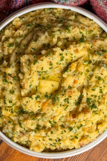 A bowl of scottish clapshot topped with melted butter and finely chopped chives, placed on a wooden surface next to a colourful, textured cloth.