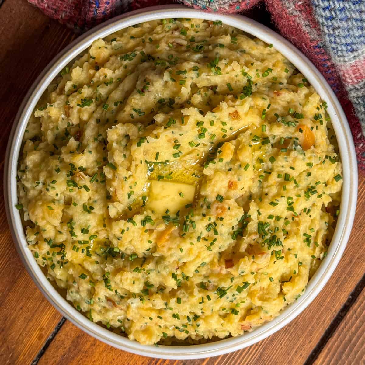 A bowl of scottish clapshot topped with melted butter and finely chopped chives, placed on a wooden surface next to a colourful, textured cloth.