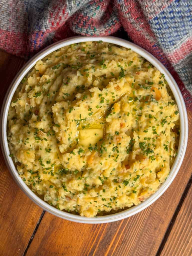 A bowl of scottish clapshot topped with melted butter and chopped chives sits on a wooden table, next to a checked cloth.