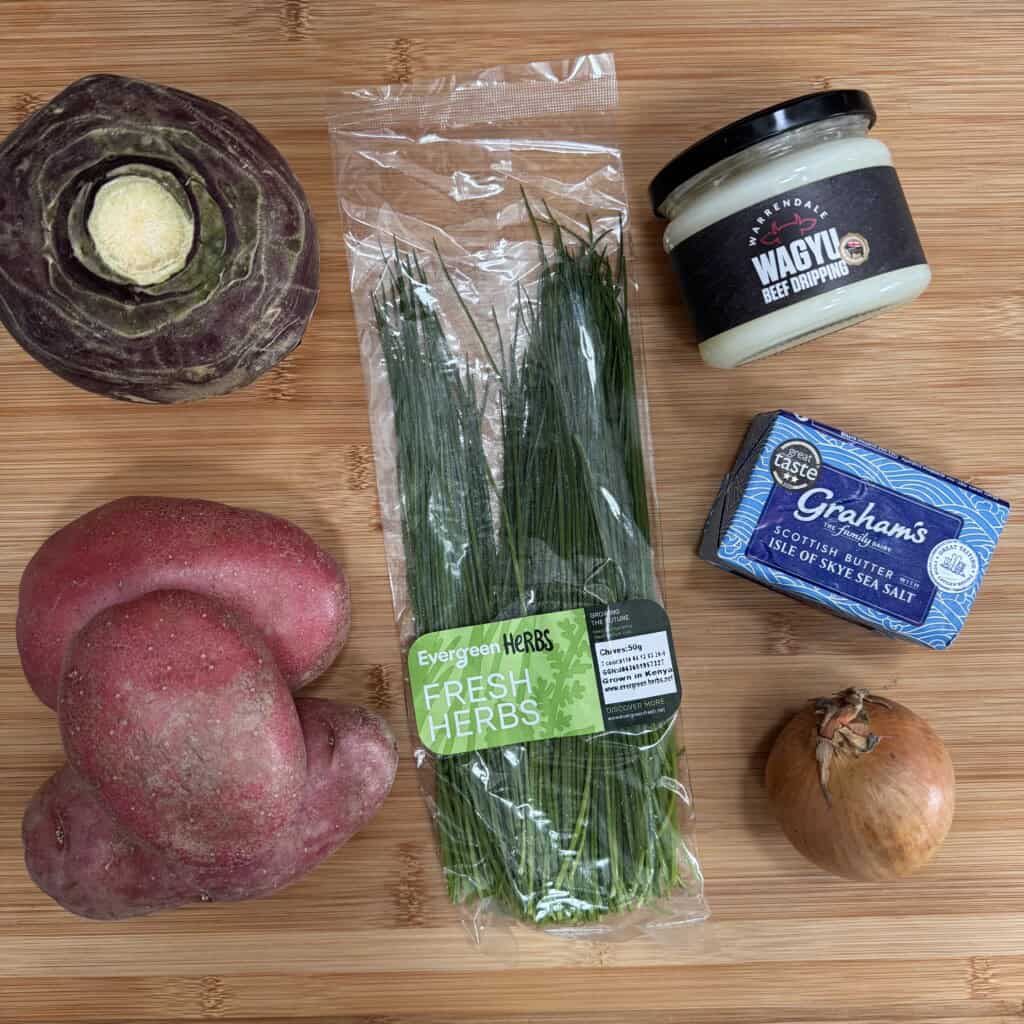 A top-down view of a wooden surface with a kohlrabi, two red potatoes, a pack of fresh chives, a jar of Wagyu beef dripping, a block of Scottish butter, and a brown onion arranged neatly.