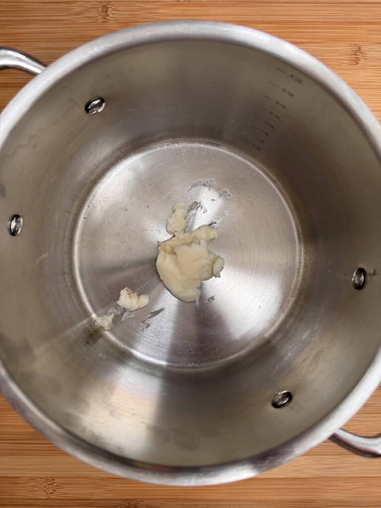 A small amount of light-coloured butter or fat is melting at the bottom of a large, empty stainless steel saucepan placed on a wooden surface.