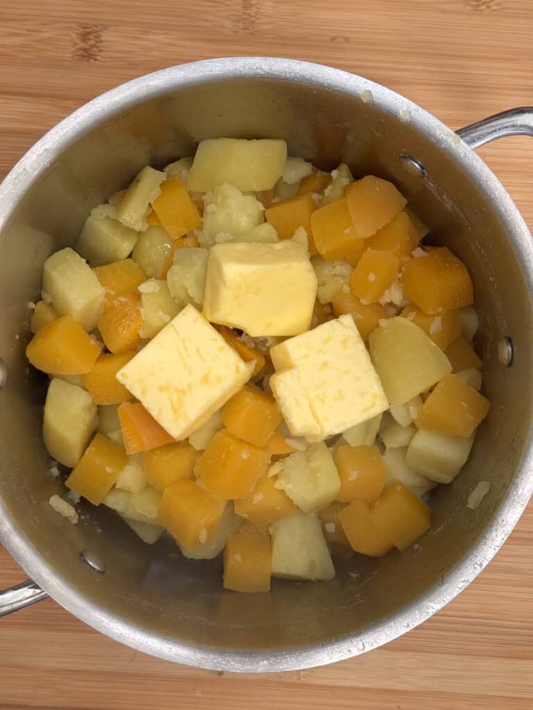A metal saucepan containing chunks of yellow and orange vegetables, likely potatoes and squash, with several cubes of butter on top, sitting on a wooden surface.