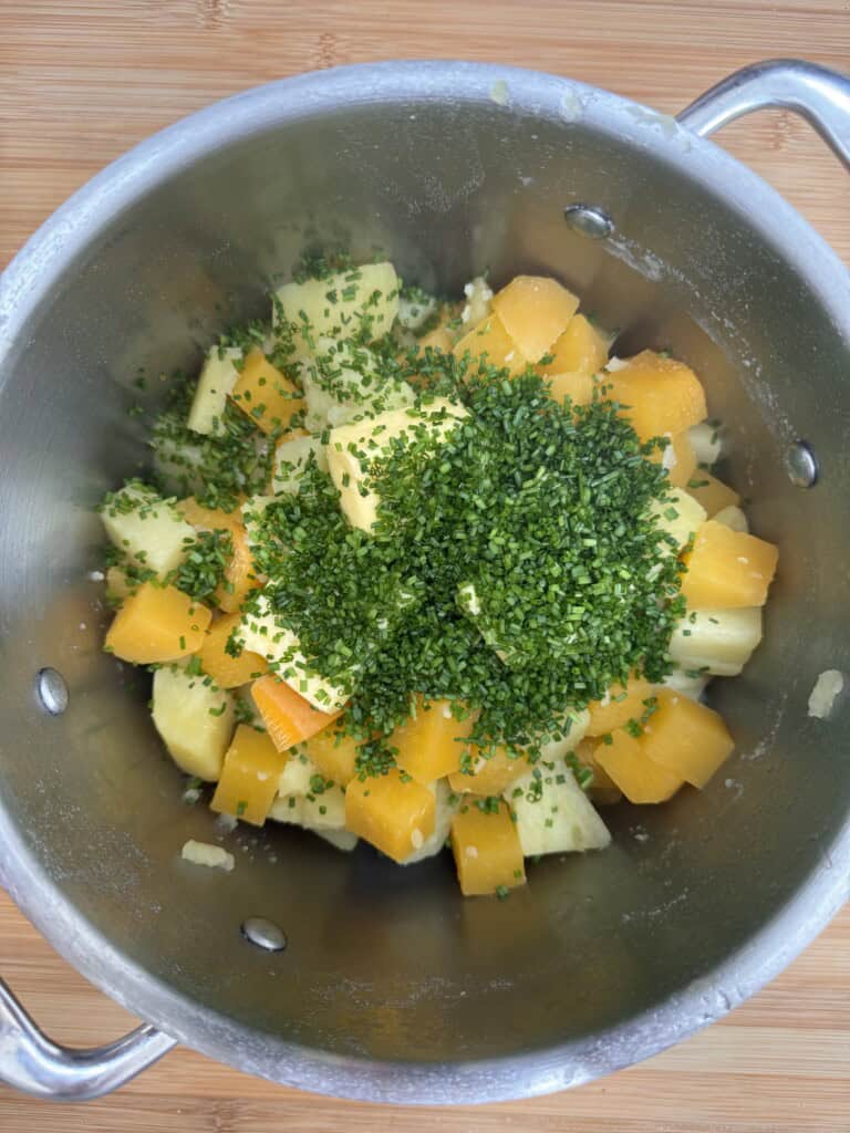 A stainless steel saucepan containing chopped yellow potatoes, orange vegetables, and a generous amount of chopped fresh green herbs, sitting on a wooden surface.