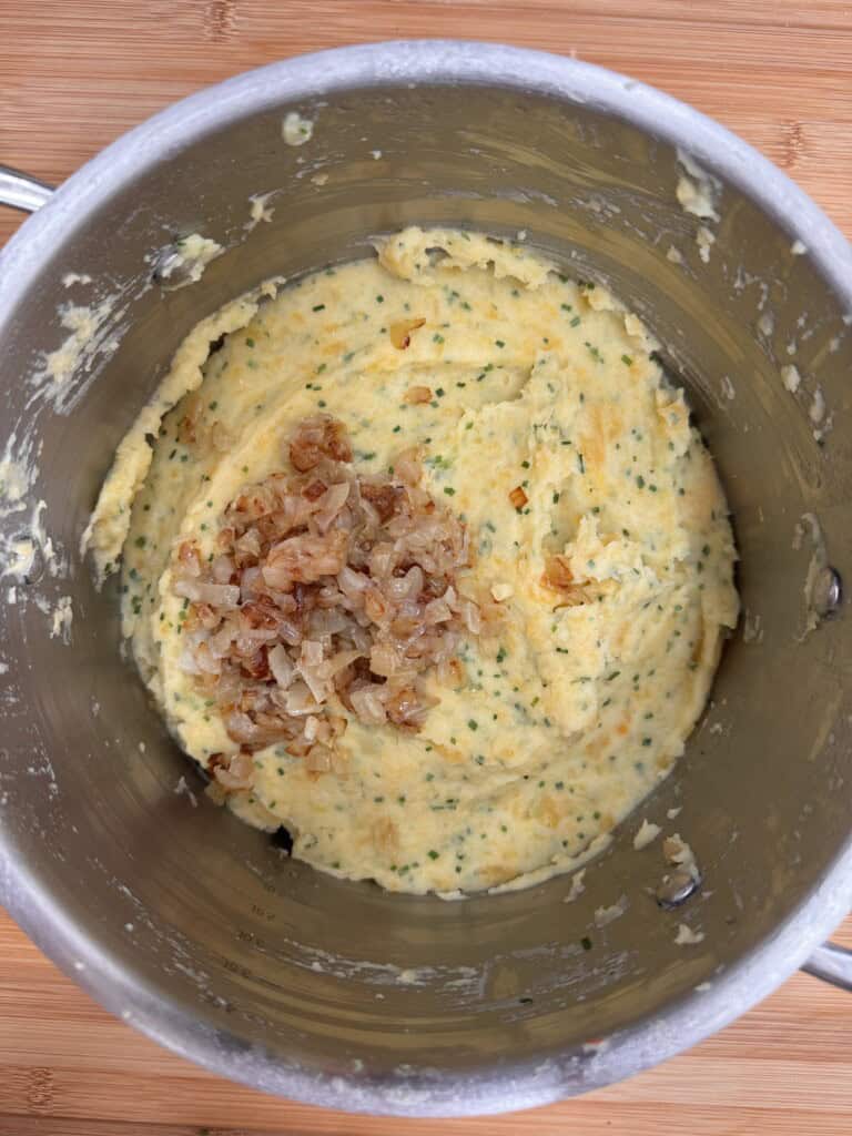 A metal saucepan containing mashed potatoes mixed with herbs, with a small pile of sautéed onions on top, sitting on a wooden surface.