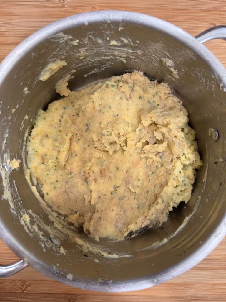 A stainless steel saucepan containing a thick, yellowish dough mixed with herbs and small vegetable pieces sits on a wooden surface. The dough has a slightly rough texture and is spread unevenly in the saucepan.