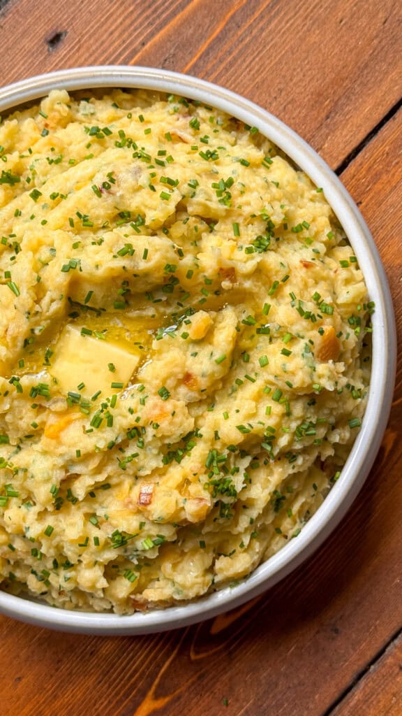 A bowl of creamy mashed potatoes topped with chopped chives and a knob of melting butter, set on a wooden table.