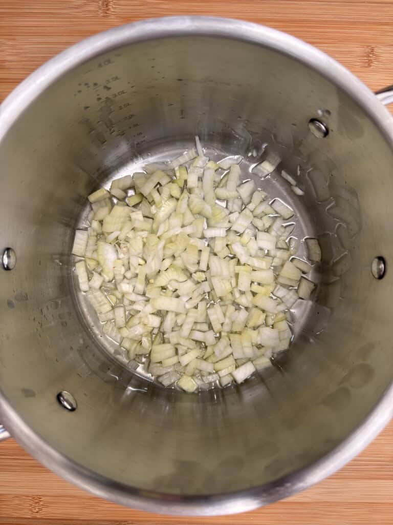 Diced onions are sautéing in oil at the bottom of a large stainless steel saucepan on a wooden surface.