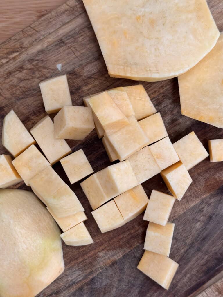 Cubed pieces of peeled butternut squash arranged on a wooden chopping board, with some larger uncut sections visible in the background.