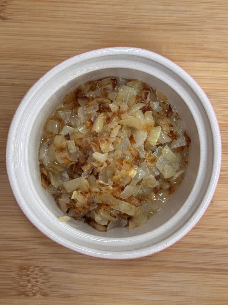 A white bowl filled with chopped, golden-brown caramelised onions sits on a wooden surface.