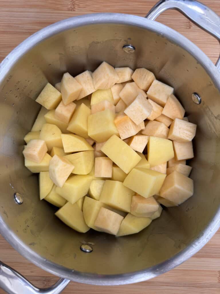 A stainless steel saucepan filled with cubed pieces of raw yellow and light orange root vegetables, likely potatoes and swedes, sitting on a wooden surface.