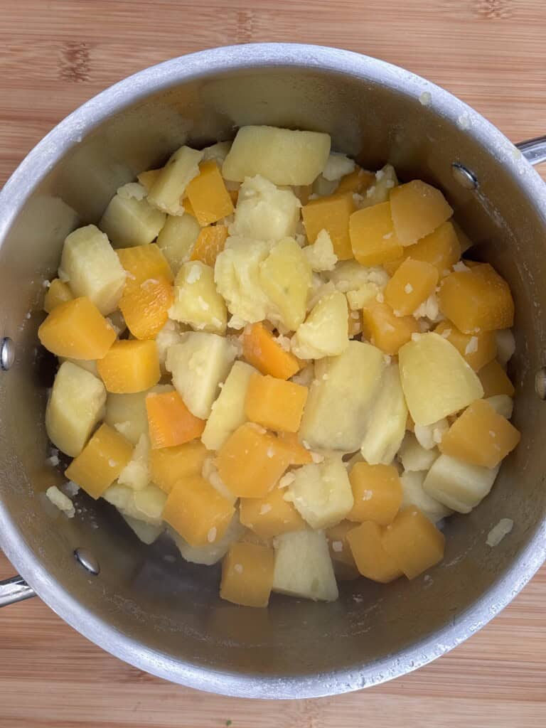 A stainless steel saucepan containing steamed, cubed yellow and white root vegetables, possibly sweet potatoes and yams, on a wooden surface.