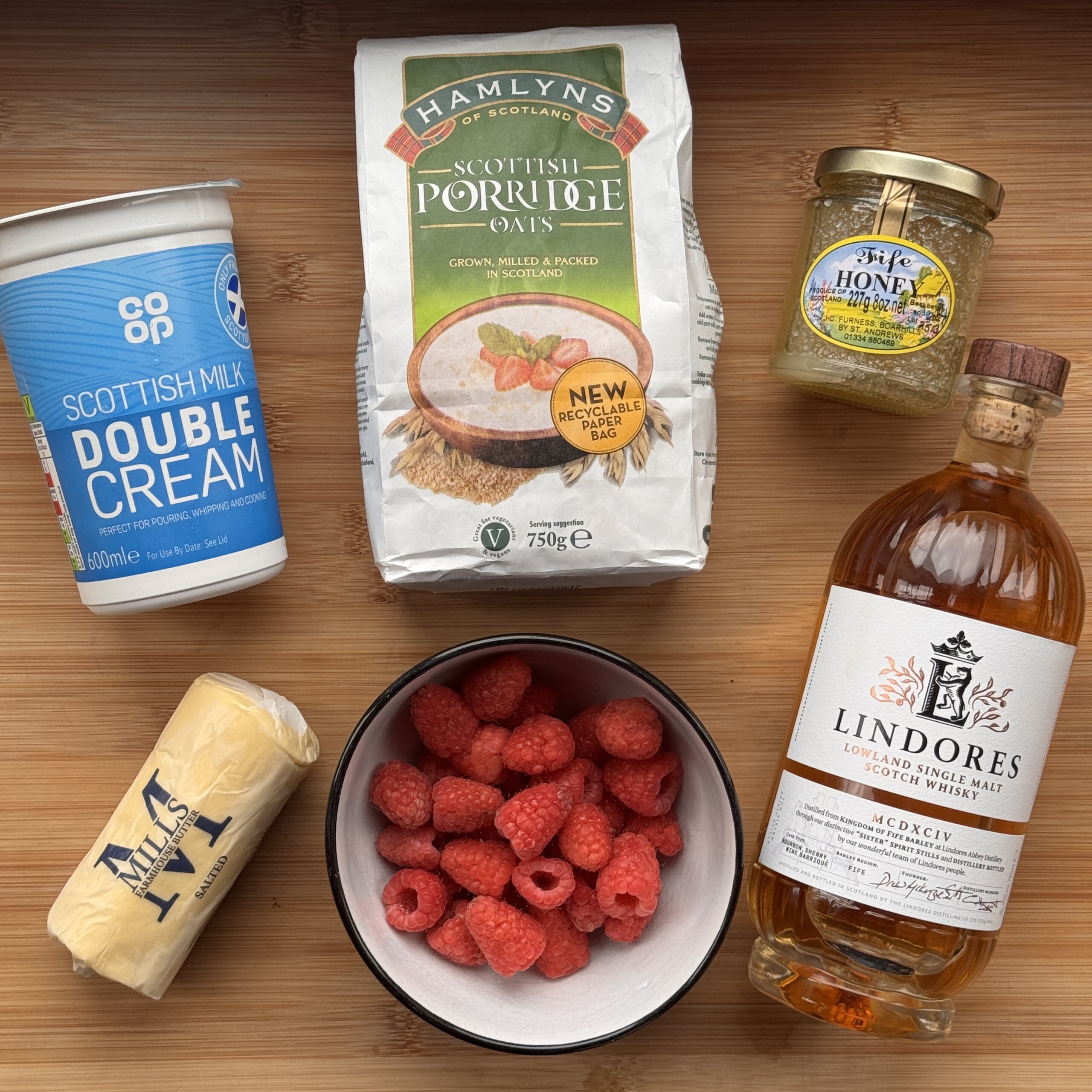 A flat lay of Scottish ingredients for Cranachan on a wooden surface: double cream, porridge oats, honey, butter, fresh raspberries in a bowl, and a bottle of Lindores whisky.