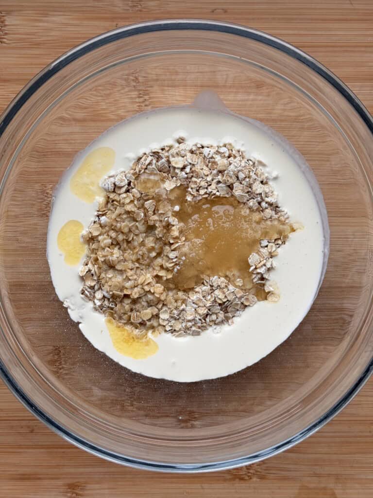 A glass bowl on a wooden surface contains oats, honey, melted butter, and cream, ready to be mixed.