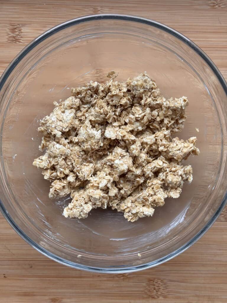 A clear glass bowl containing a mixture of oats and what appears to be a sticky or dough-like substance, sitting on a wooden surface.