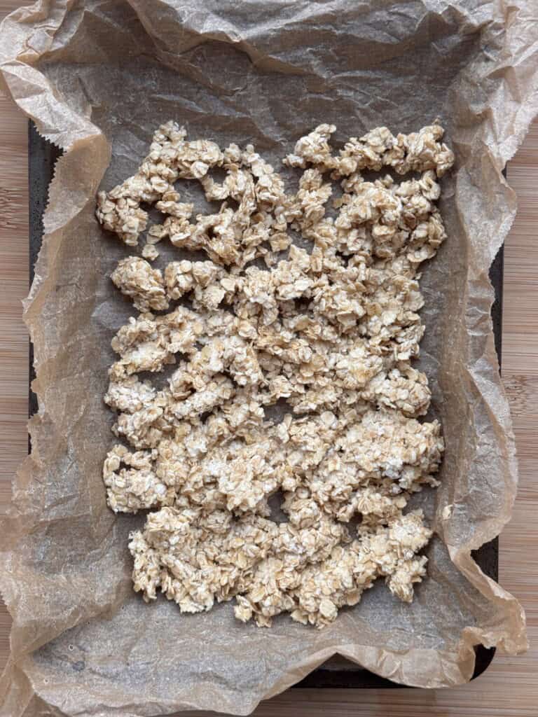 A baking tray lined with brown greaseproof paper holds an uneven, flattened layer of oat mixture, likely prepared for baking, on a wooden surface.