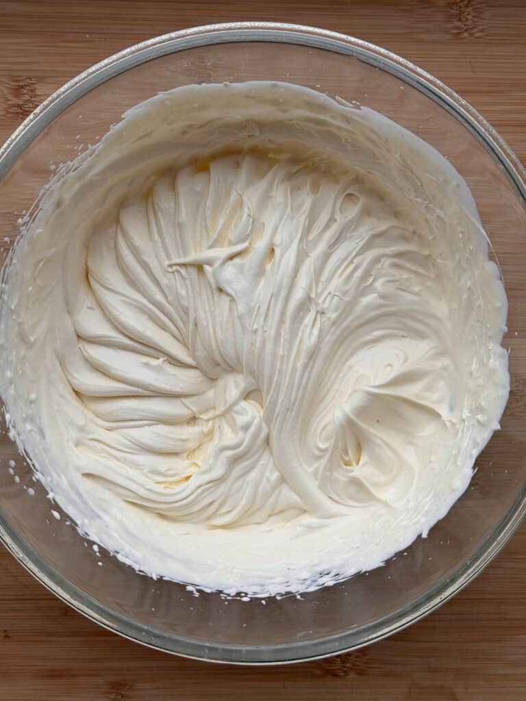 A glass bowl filled with creamy, whipped mixture with soft peaks and swirled patterns on a wooden surface.