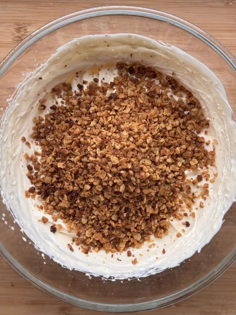 A glass bowl filled with creamy white mixture topped with a generous layer of finely chopped toasted nuts, sitting on a wooden surface.