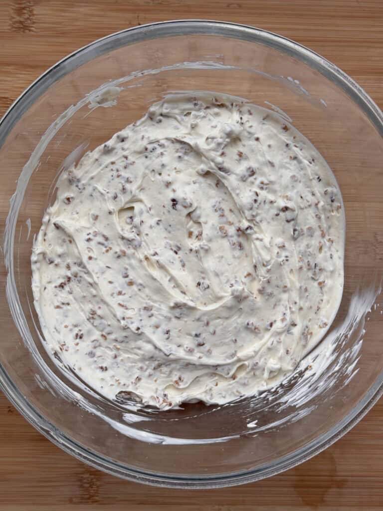 A glass bowl containing a creamy, speckled mixture sits on a wooden surface. The mixture appears to have small brown seeds or grains evenly distributed throughout.