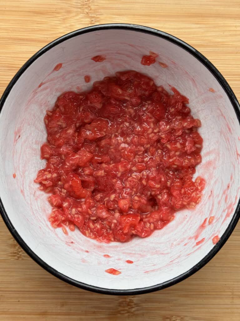 A close-up of a bowl filled with mashed raspberries on a light wooden surface. The bright red fruit is crushed, showing a juicy and textured mixture inside a white bowl with a dark rim.