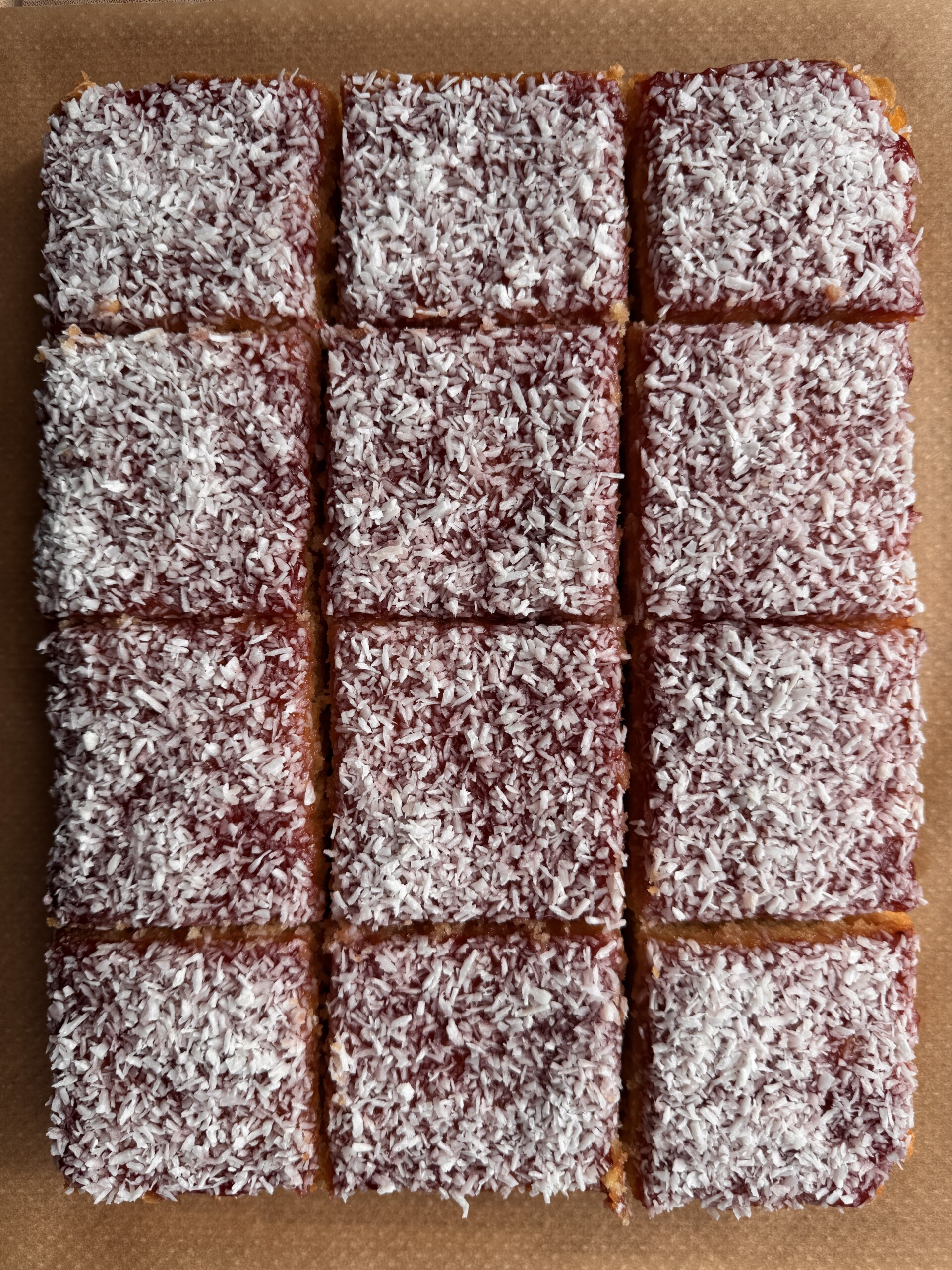 Twelve neatly cut squares of jam and coconut sponge topped with a layer of red jam and sprinkled with desiccated coconut, arranged on a brown surface.