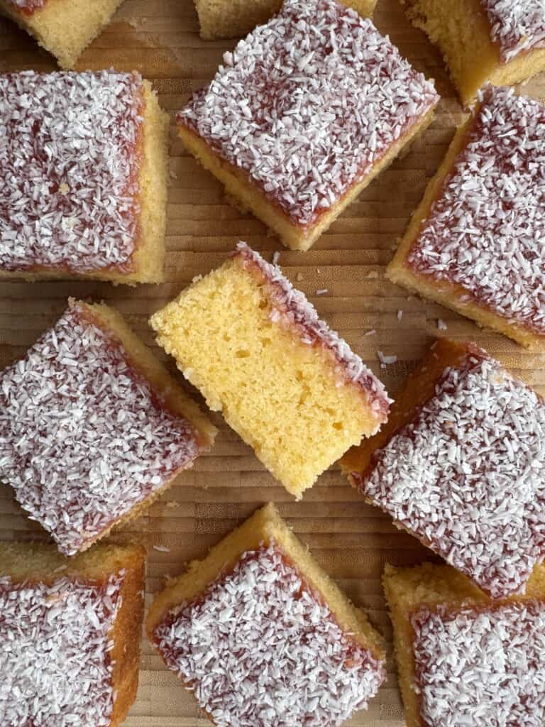 Several pieces of jam and coconut sponge are topped with a layer of red jam and sprinkled with desiccated coconut, arranged closely together on a wooden surface.