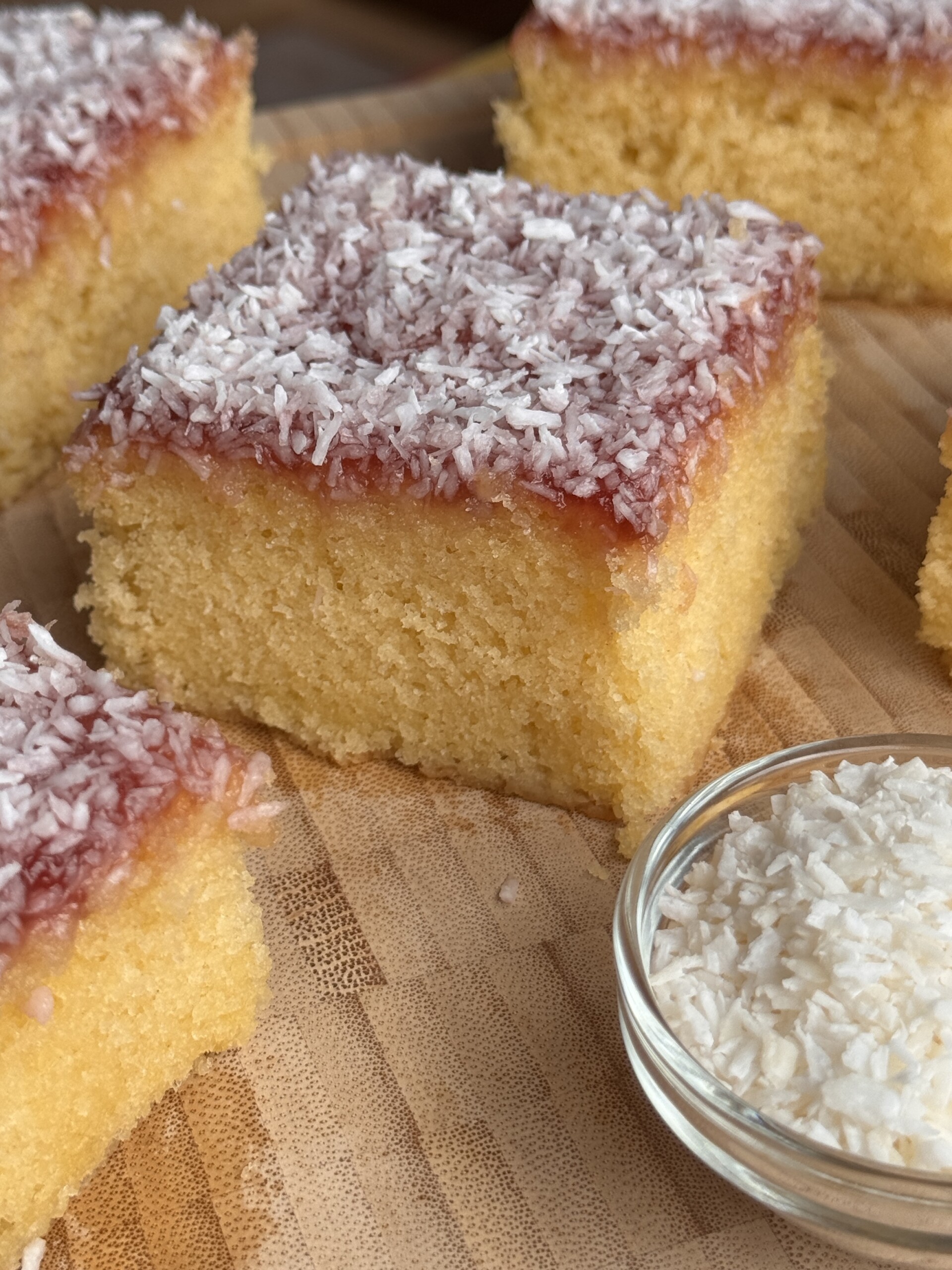 A close-up of square pieces of jam and coconut sponge topped with pink icing and desiccated coconut, arranged on a wooden surface. A small glass bowl filled with desiccated coconut is beside the cake slices.