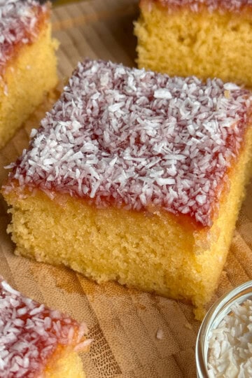 Squares of jam and coconut sponge topped with a layer of red jam and sprinkled with desiccated coconut, arranged on a wooden board, with a small dish of coconut nearby.