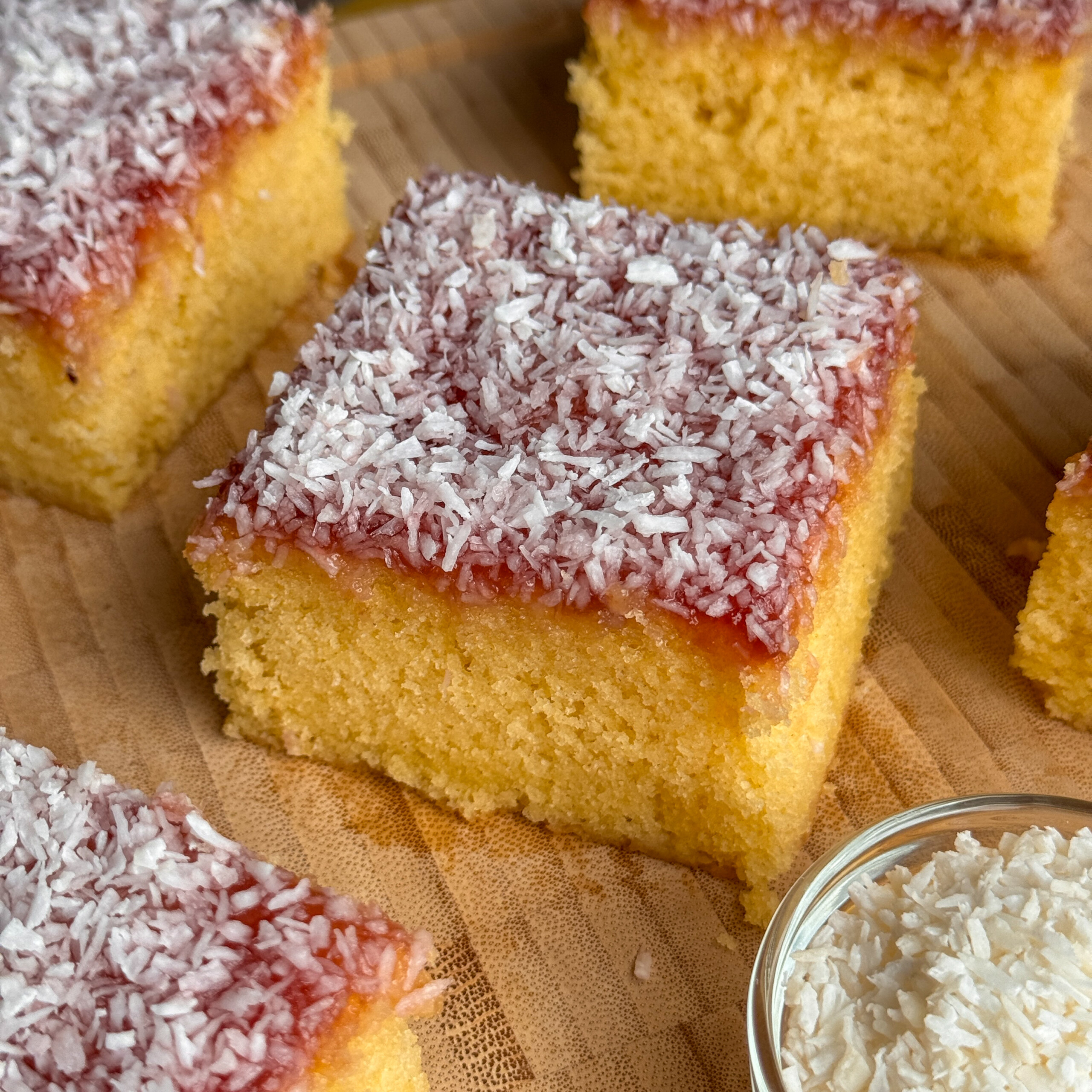 Squares of jam and coconut sponge topped with a layer of red jam and sprinkled with desiccated coconut, arranged on a wooden board, with a small dish of coconut nearby.
