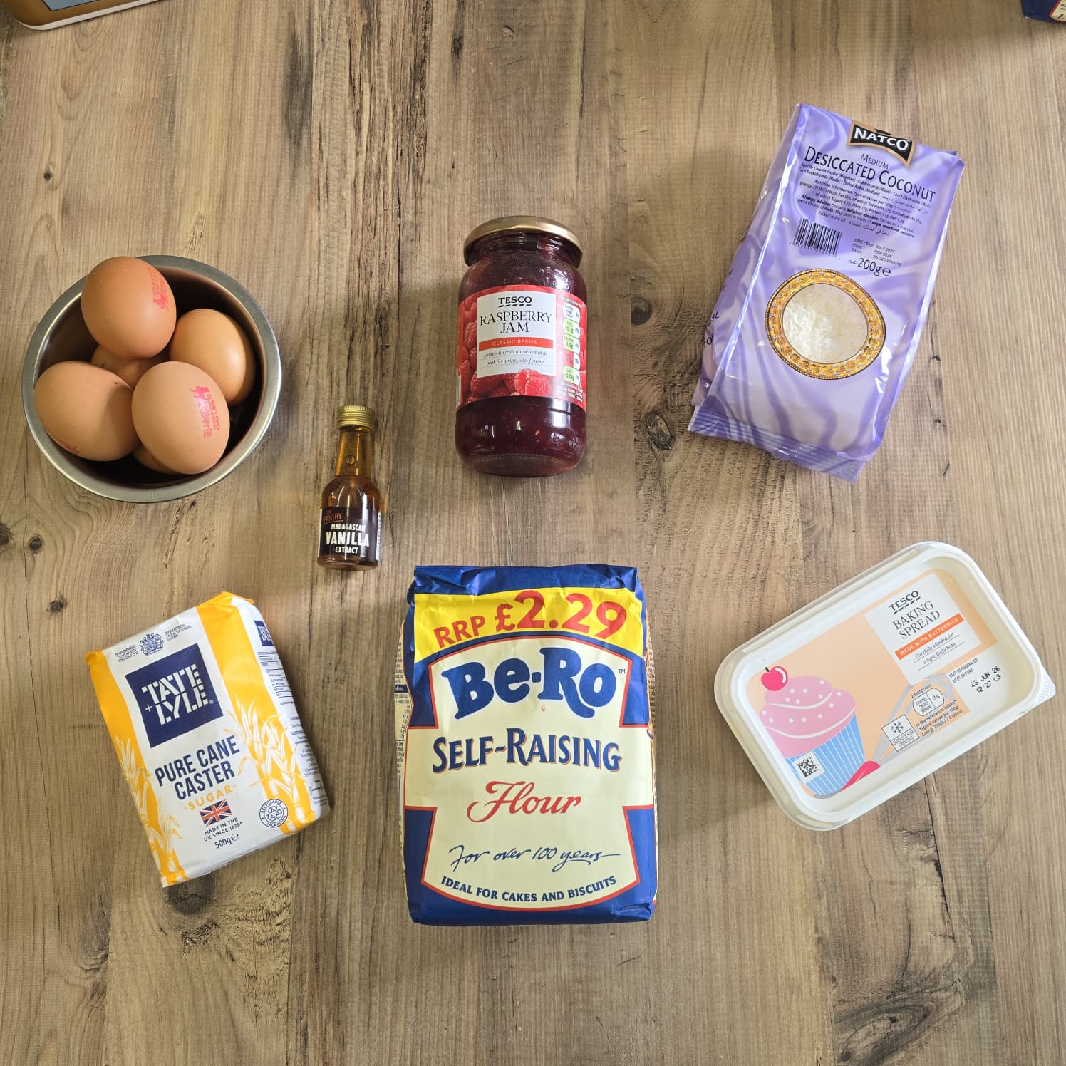 Baking ingredients for jam and coconut sponge on a wooden surface: a bowl of eggs, a small bottle of vanilla, caster sugar, self-raising flour, strawberry jam, desiccated coconut, and a tub of spreadable butter.