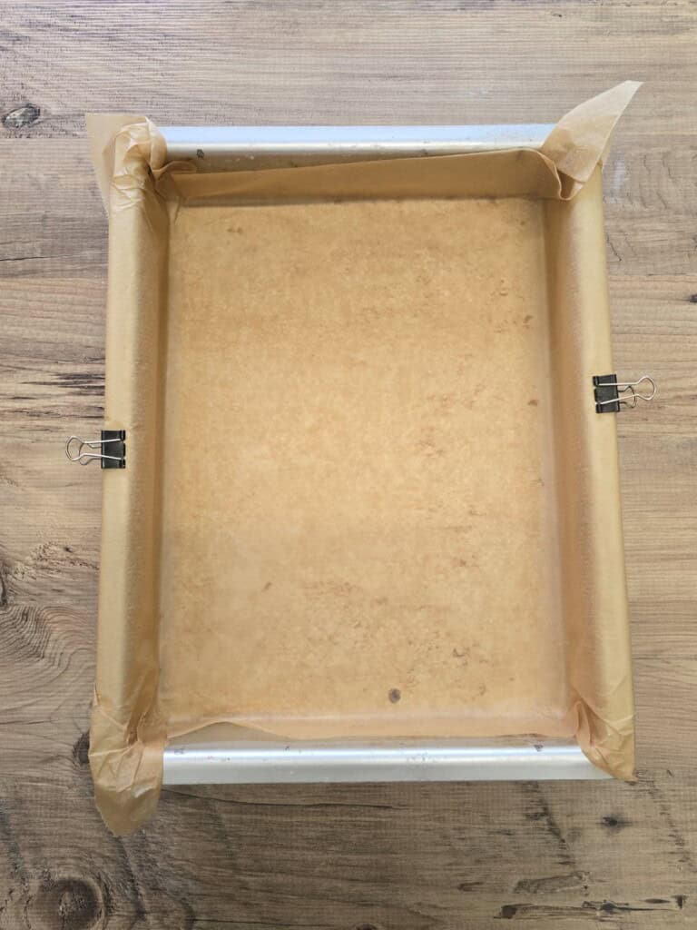 A rectangular baking tray lined with greaseproof paper is secured at the sides with two metal bulldog clips, placed on a wooden surface.