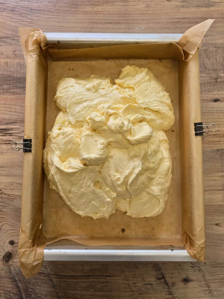 A parchment-lined rectangular baking tin with yellow cake mixture spread unevenly inside, secured with bulldog clips, sits on a wooden surface.