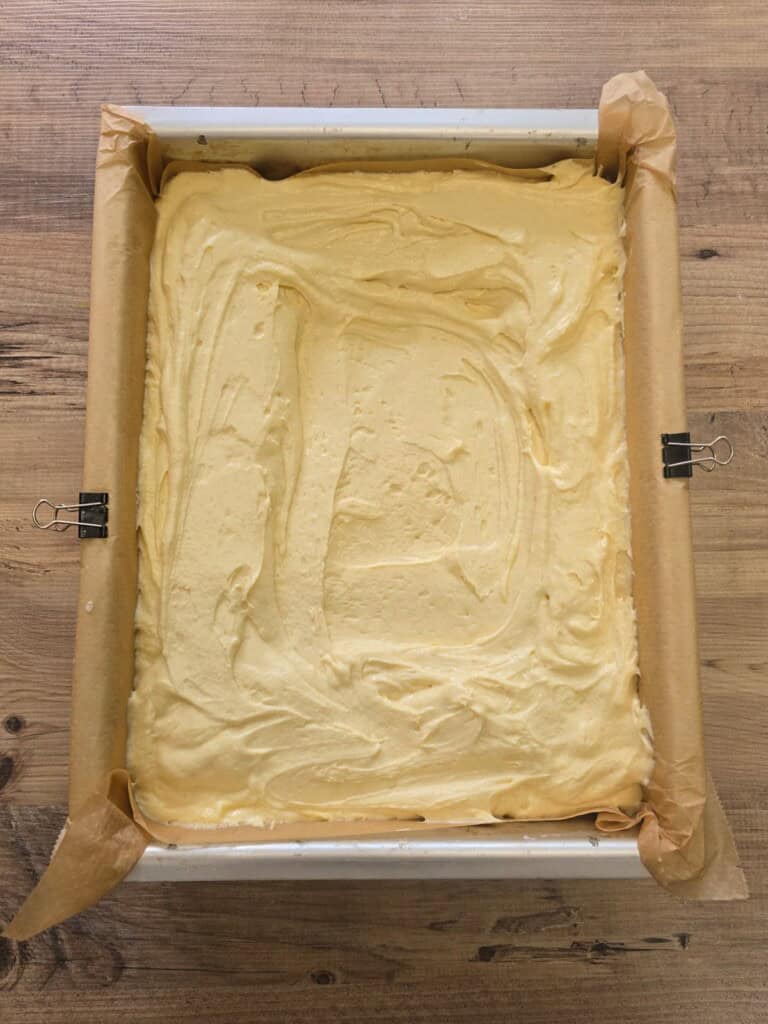 A square baking tin lined with greaseproof paper and filled with smooth, pale yellow cake mixture, secured with metal bulldog clips, sits on a wooden surface.