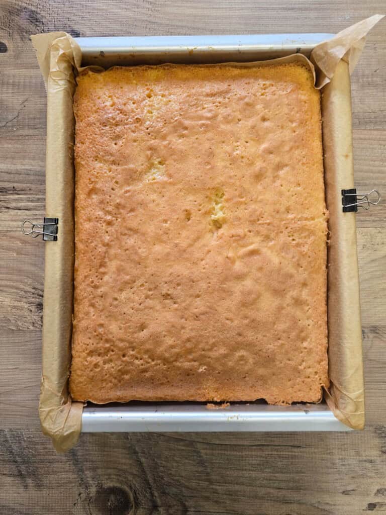 A freshly baked golden sponge cake in a square tin lined with baking paper, held at the sides with metal clips, sits on a wooden surface.