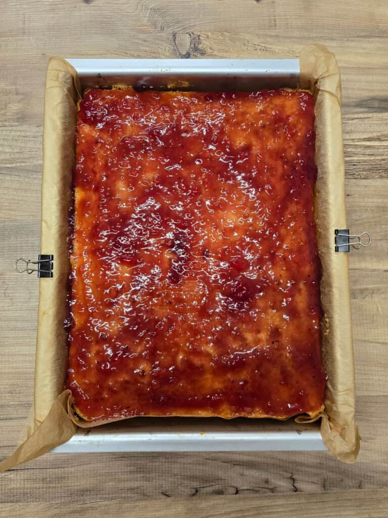 A rectangular tray lined with baking parchment holds a sheet cake topped with a glossy layer of red jam or fruit glaze, sitting on a wooden surface. The cake is uncut and appears freshly baked.