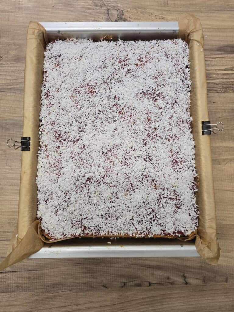 A rectangular tray lined with brown baking paper holds jam and coconut sponge covered with a layer of desiccated coconut. Two black bulldog clips hold the paper in place on the tray, which sits on a wooden surface.