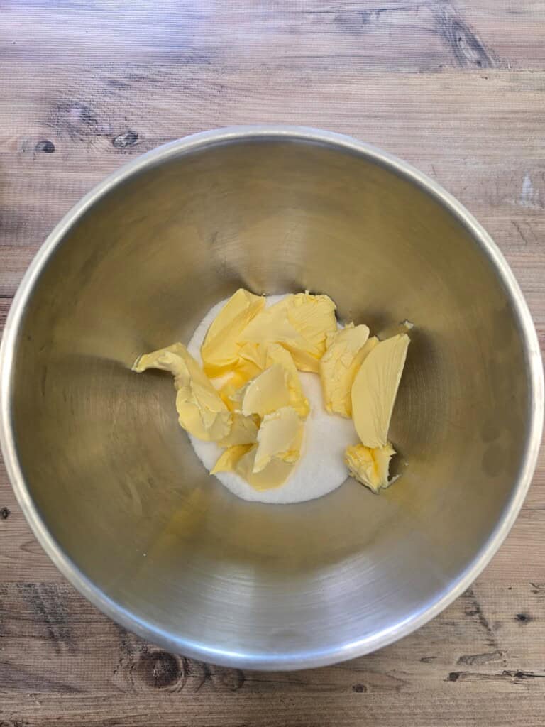 A metal mixing bowl containing a mound of caster sugar and several dollops of yellow butter, placed on a wooden surface.