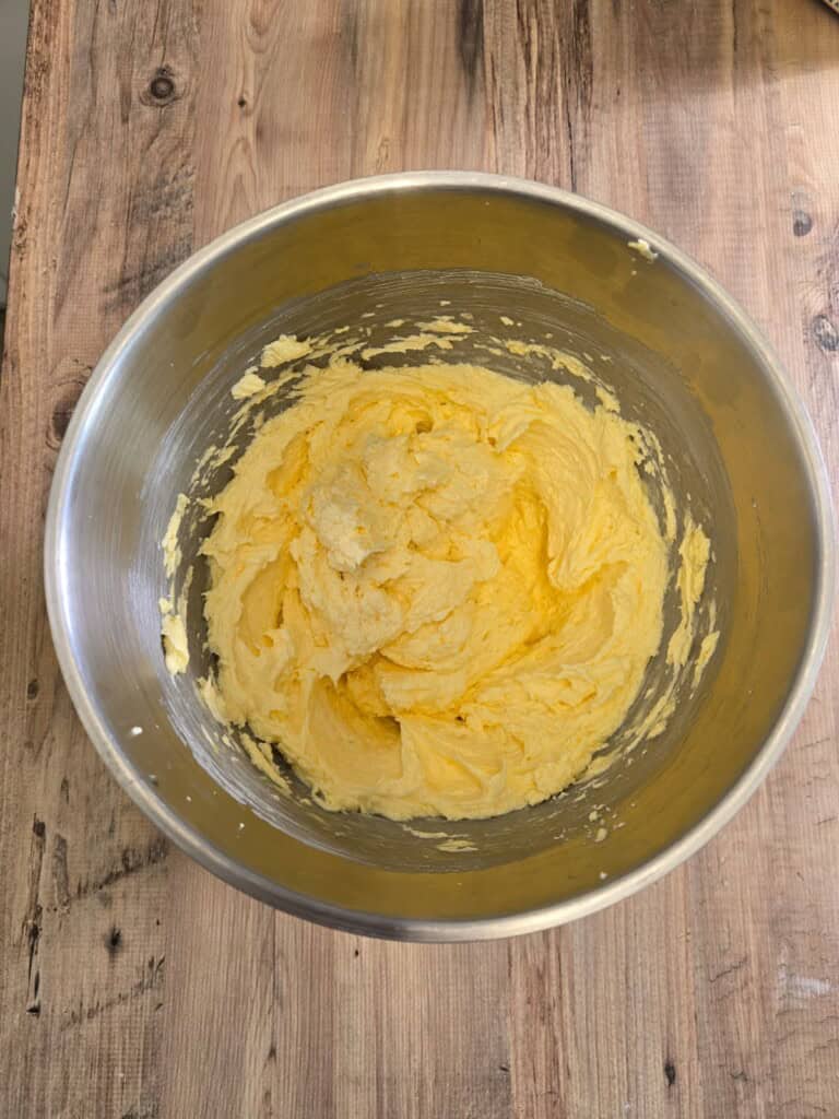 A metal mixing bowl containing a creamy yellow mixture sits on a rustic wooden surface. The mixture appears to be well-mixed and has a smooth, fluffy texture.