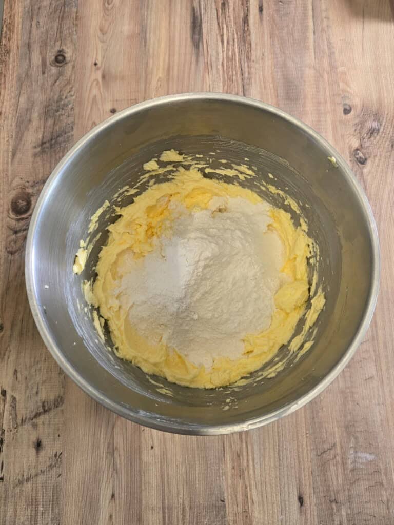 A metal mixing bowl containing creamed butter and sugar, with a mound of flour added on top, sits on a wooden surface.