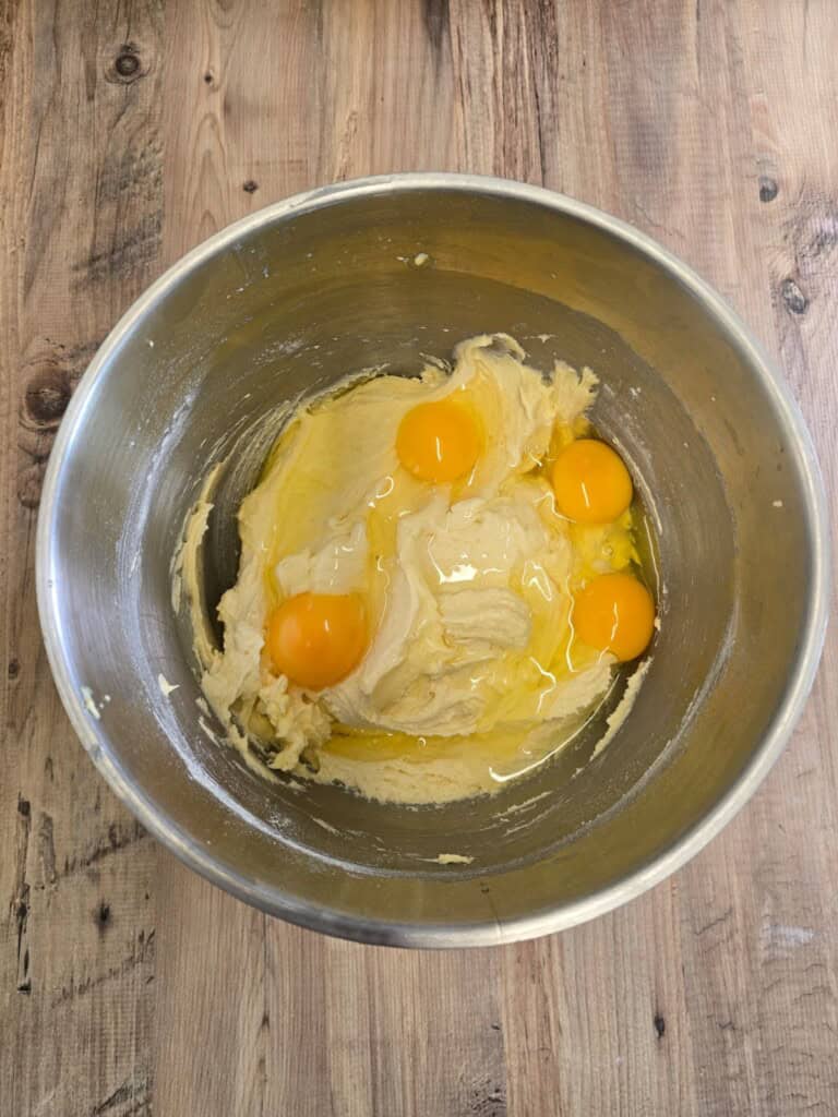 A metal mixing bowl contains creamed butter and sugar with four cracked eggs on top, all set on a wooden surface.