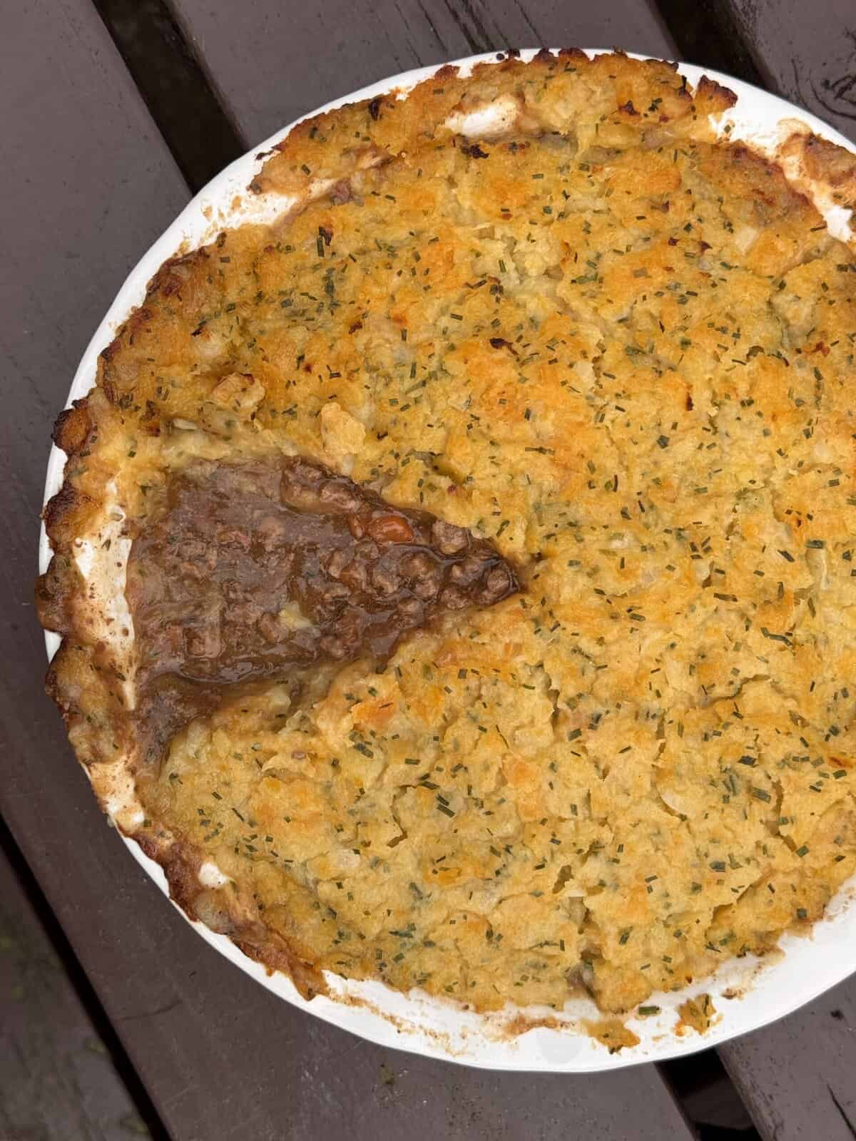 A round mince pie in a white dish with a golden-brown clapshot topping. A wedge has been served, revealing the savoury meat and vegetable filling beneath. The dish sits on a wooden surface.