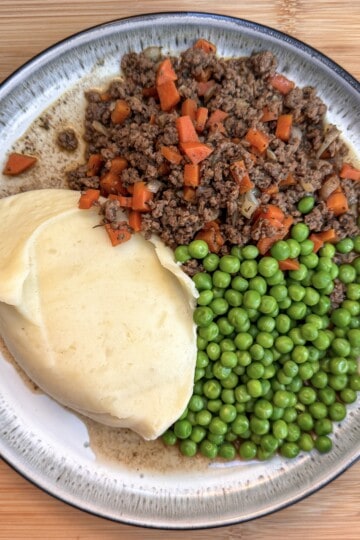 A plate of food with mince and tatties - beef mince and diced carrots, mashed potatoes, and green peas, served on a round ceramic dish over a wooden surface.