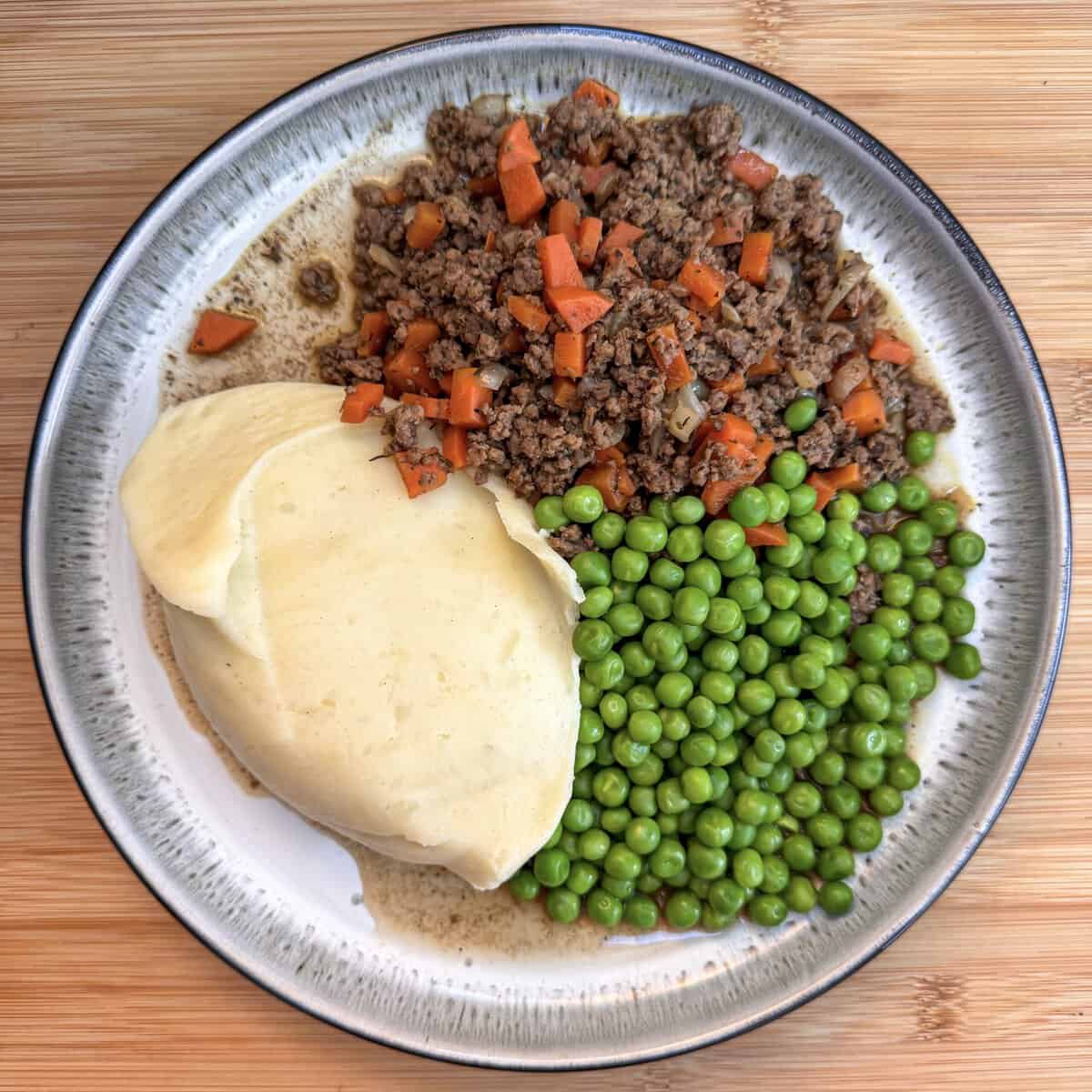 A plate of food with mince and tatties - beef mince and diced carrots, mashed potatoes, and green peas, served on a round ceramic dish over a wooden surface.
