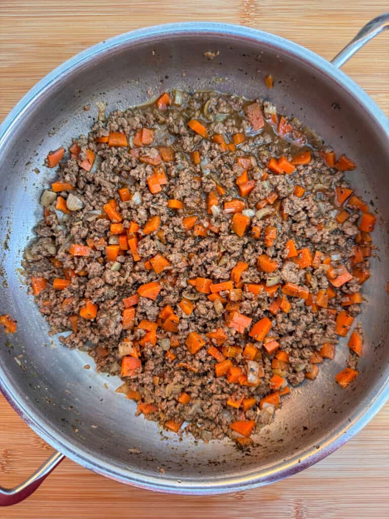 A stainless steel pan on a wooden surface contains cooked minced beef mixed with diced carrots and onions. The ingredients are evenly combined and appear to be sautéed.