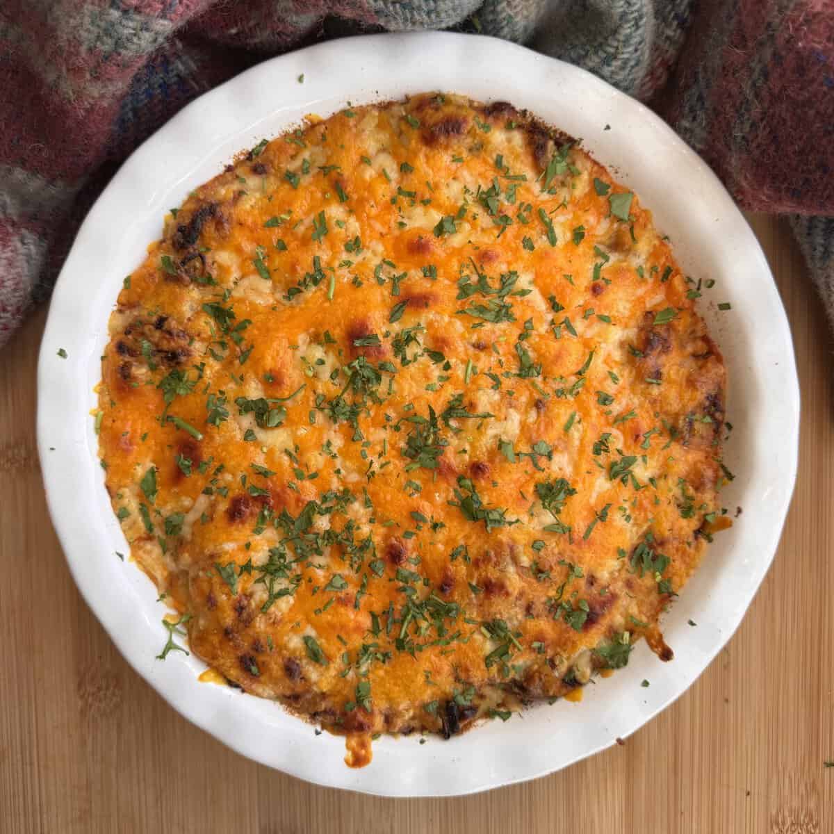 A baked casserole topped with melted golden cheese and chopped herbs, served in a white dish on a wooden surface with a checked cloth in the background.