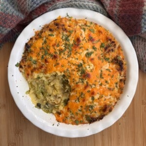 A round casserole dish filled with shepherd’s pie topped with golden mashed potatoes, parsley, and a portion scooped out, revealing a vegetable filling. The dish is placed on a wooden surface next to a patterned cloth.