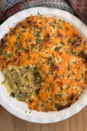 A round casserole dish filled with shepherd’s pie topped with golden mashed potatoes, parsley, and a portion scooped out, revealing a vegetable filling. The dish is placed on a wooden surface next to a patterned cloth.