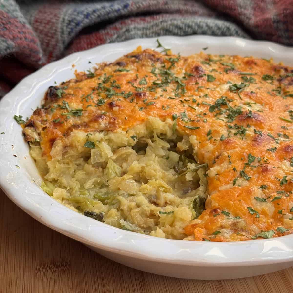A classic rumbledethumps with a golden, cheesy crust and fresh green herbs on top sits in a white dish. A serving reveals its creamy, vegetable-filled interior, while a tartan cloth adds cosy charm in the background.