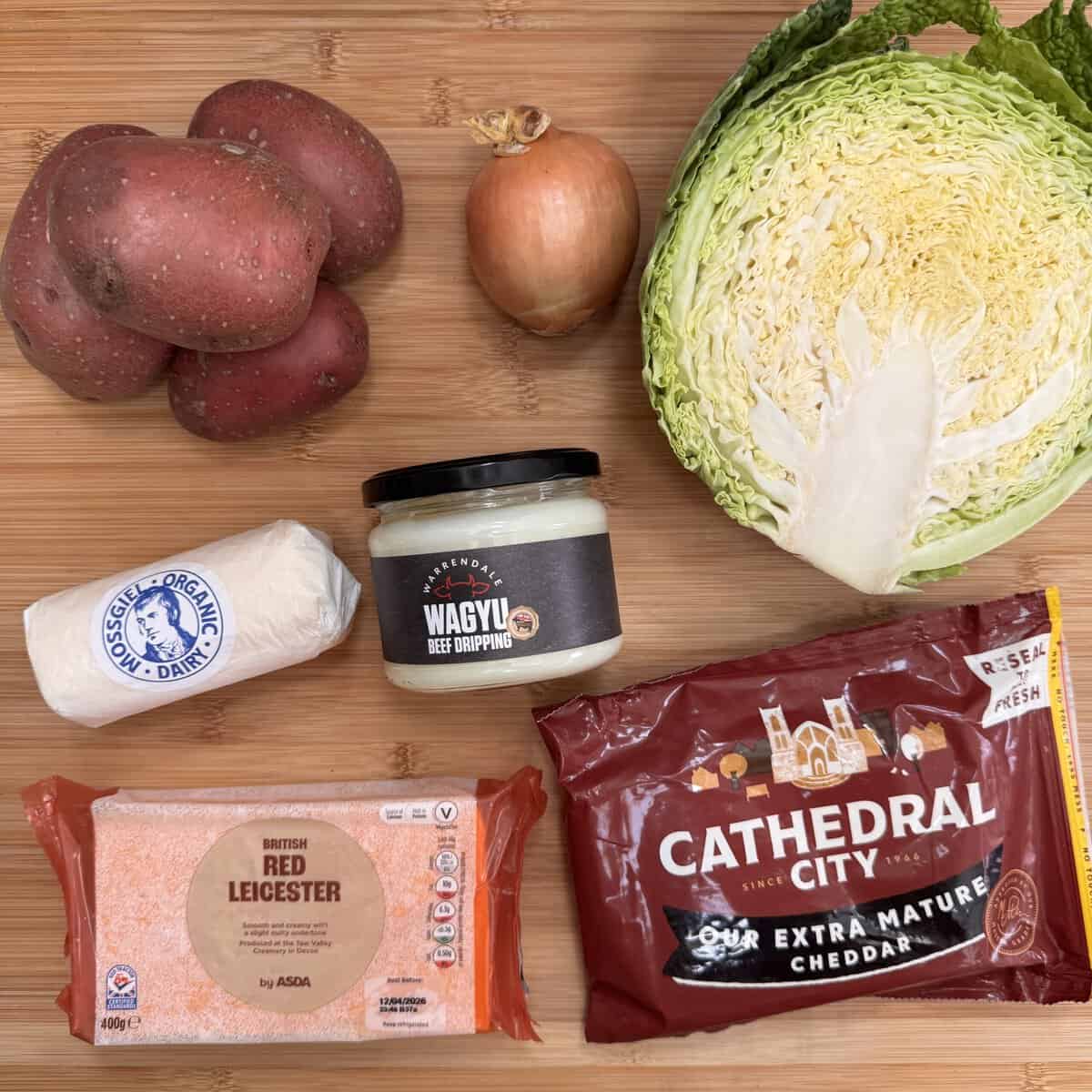 A wooden surface displaying ingredients for rumbledethumps: red potatoes, a brown onion, half a green cabbage, a log of goat's cheese, Wagyu beef dripping, Red Leicester cheese, and Cathedral City cheddar.